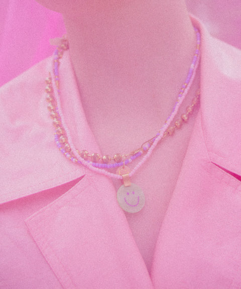 A person wears a pink blazer and layered classic necklaces, including the WALD Berlin Pop Smilie Medallion Peach, shown from the shoulders up.