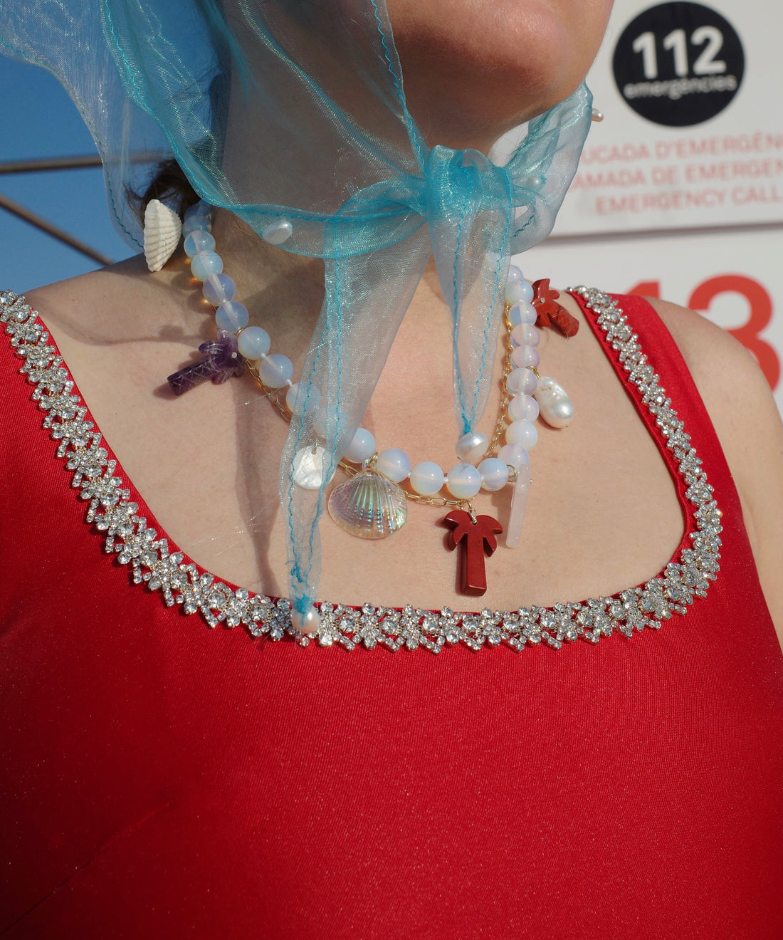 A person in a red dress with a jeweled neckline and layered necklaces—one is the Mexicana La Bamba Necklace by WALD Berlin—wears a blue sheer scarf tied under the chin. An emergency sign appears in the background.