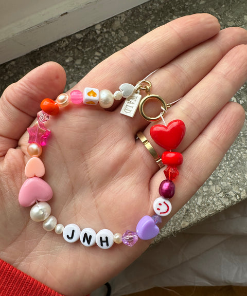 A hand holds the WALD Berlin Personalized bracelet, featuring colorful beads, sweetwater pearls, heart-shaped accents, a red heart charm, and custom letter beads spelling “JNH.”.