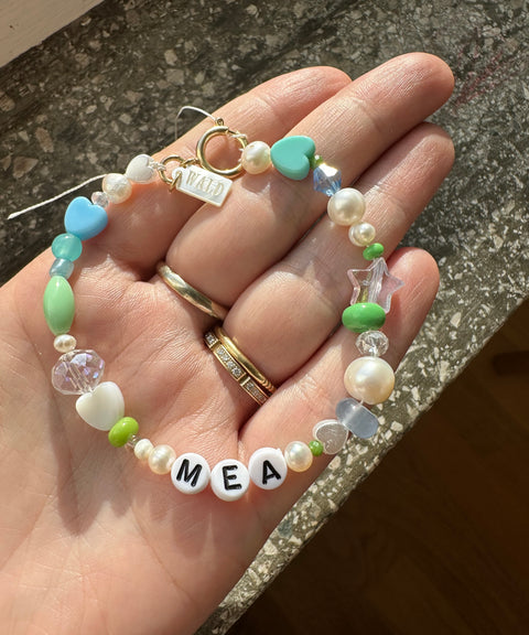 A hand holds a WALD Berlin personalized bracelet featuring the letters “MEA,” heart and star-shaped beads, sweetwater pearls, green accents, and a clasp adorned with a small charm.