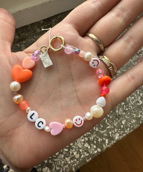 A hand holding the WALD Berlin Personalized bracelet, featuring colorful beads with hearts, sweetwater pearls, smiley faces, and the letters “L” and “C.”.