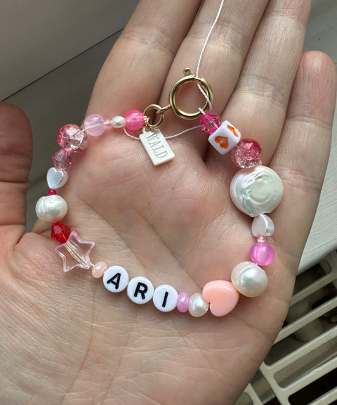 A hand holds the WALD Berlin personalized bracelet, featuring pink, white, and red beads with sweetwater pearls, a gold clasp, a “WILD” charm, and white letter beads spelling “ARI.”.