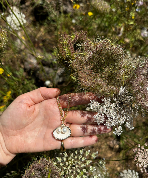 A hand holds the WALD Berlin Drop It Like It’s Hot Shell Necklace, featuring a shell and two pearls, against a backdrop of blooming wildflowers and Queen Anne's lace outdoors.