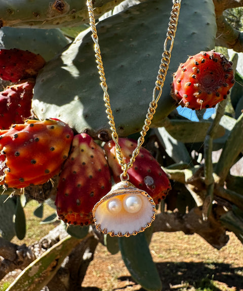 The Drop It Like It’s Hot Shell Necklace by WALD Berlin features a gold-plated chain, shell pendant, and two pearls, shown hanging before a prickly pear cactus with red fruit—a striking piece of handmade jewelry.