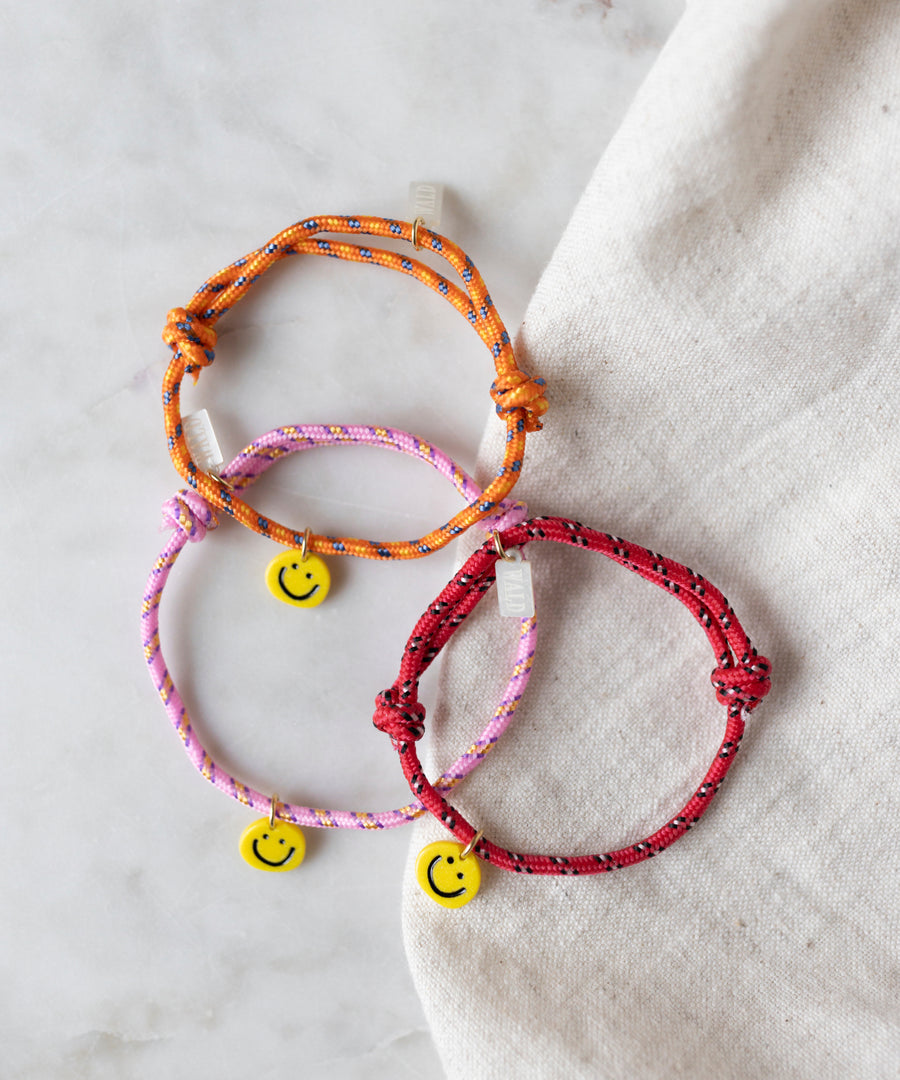 The WALD Berlin Smilie Dude Orange Friendship Bracelet, along with red and pink woven rope bracelets featuring small yellow smiley face charms, are displayed on a light fabric background.