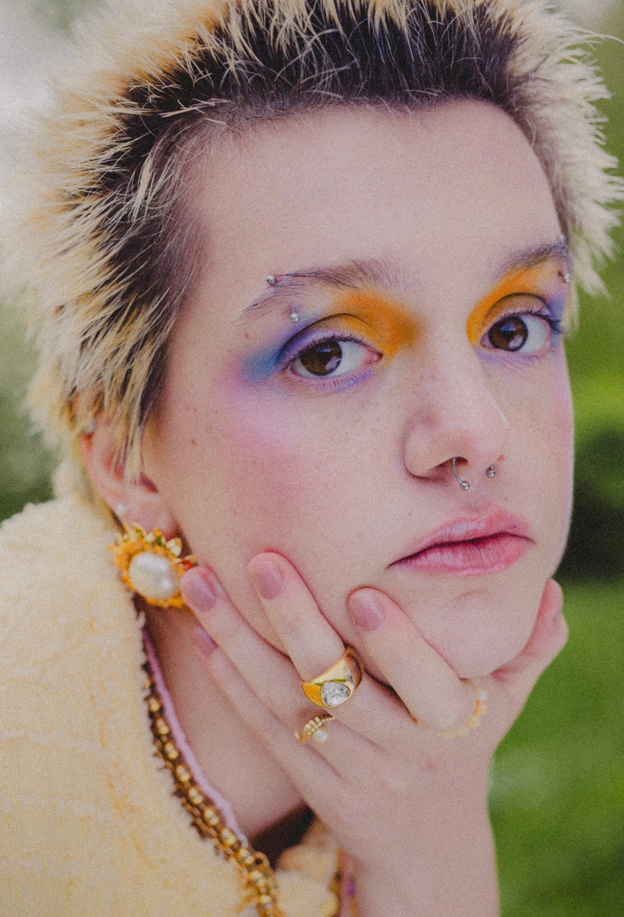 A person with short spiked hair and colorful eye makeup rests their chin on their hand, wearing WALD Berlin's SAMPLE SALE Pop Soleil Earrings, gold plated statement jewelry, set against a blurred outdoor background.