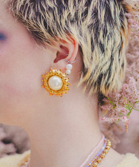 Close-up of a person with short, bleached and dark hair wearing WALD Berlin's SAMPLE SALE Pop Soleil Earrings—bold statement jewelry styled with a pearl ear cuff and layered beaded necklaces.