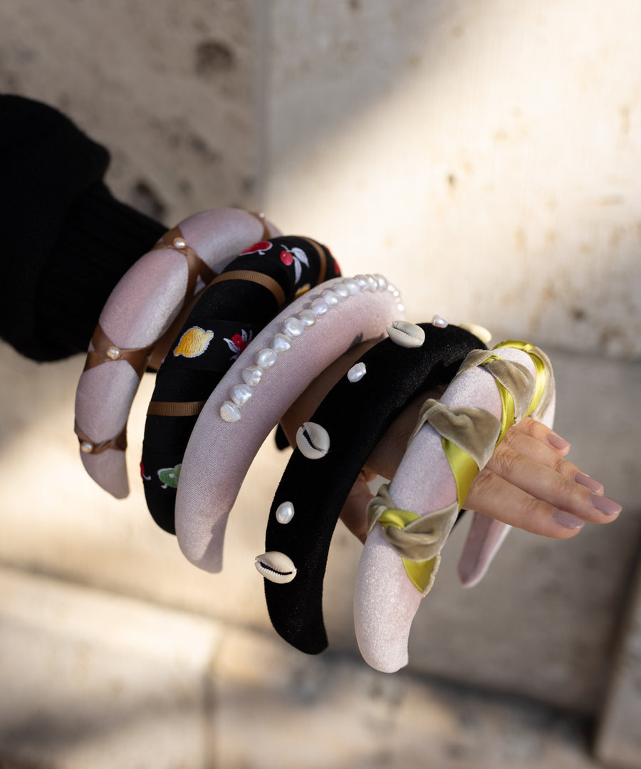 A person’s arm shows off several padded WALD Berlin headbands, like the SAMPLE SALE Simone de Beauvoir Black Headband, in unique designs—pearls, embroidery, shells, and ribbons—against a blurred stone background.
