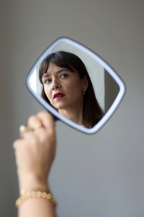 A woman with dark hair and red lipstick is reflected in a handheld mirror, holding up her hand adorned with 18k gold-plated jewelry and WALD Berlin’s Valentines Day special bow heart earrings sparkling with Swarovski stones.