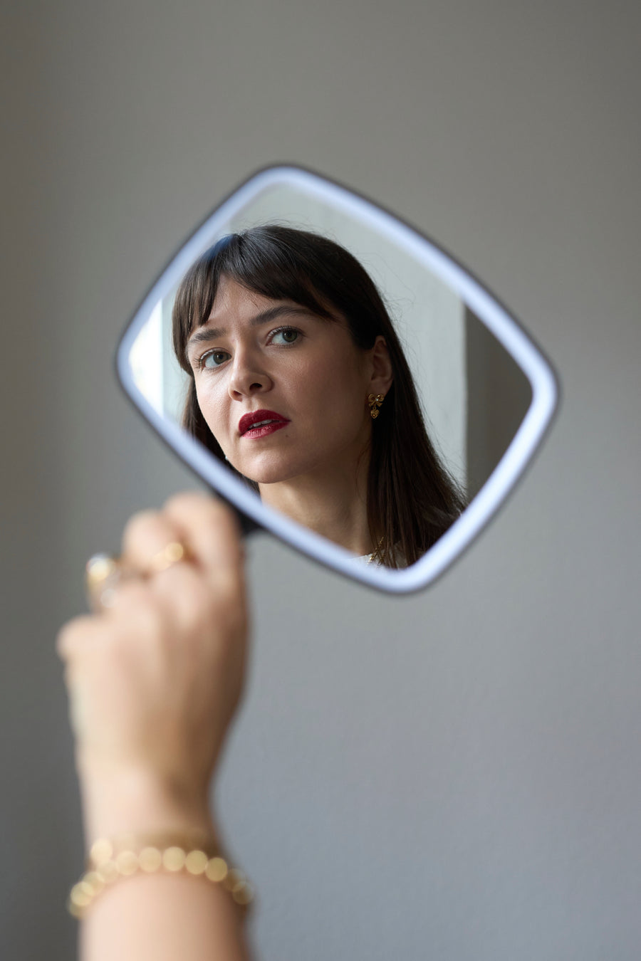 A woman with dark hair and red lipstick is reflected in a handheld mirror, holding up her hand adorned with 18k gold-plated jewelry and WALD Berlin’s Valentines Day special bow heart earrings sparkling with Swarovski stones.