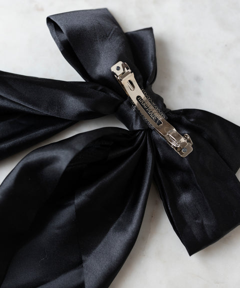The Happy Holidays Black Hair Bow by WALD Berlin, a large satin hair clip ideal for festive styling, is showcased on a white marble surface.