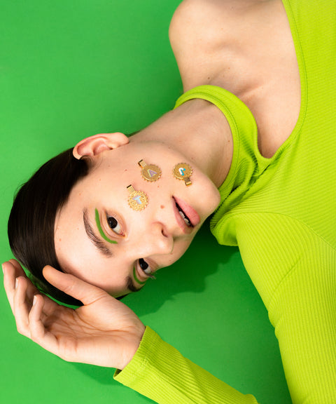 A person in a lime green top lies on a green surface with gold flower-shaped stickers, green makeup, and wears a WALD Berlin Sample saleSay My Name Gold Necklace of adjustable length around their neck.
