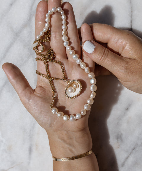 A hand displays the WALD Berlin Pas De Deux Necklace, featuring a handmade gold chain with a sweetwater pearl pendant, alongside a matching pearl bracelet on a marble surface, all complemented by white nail polish.