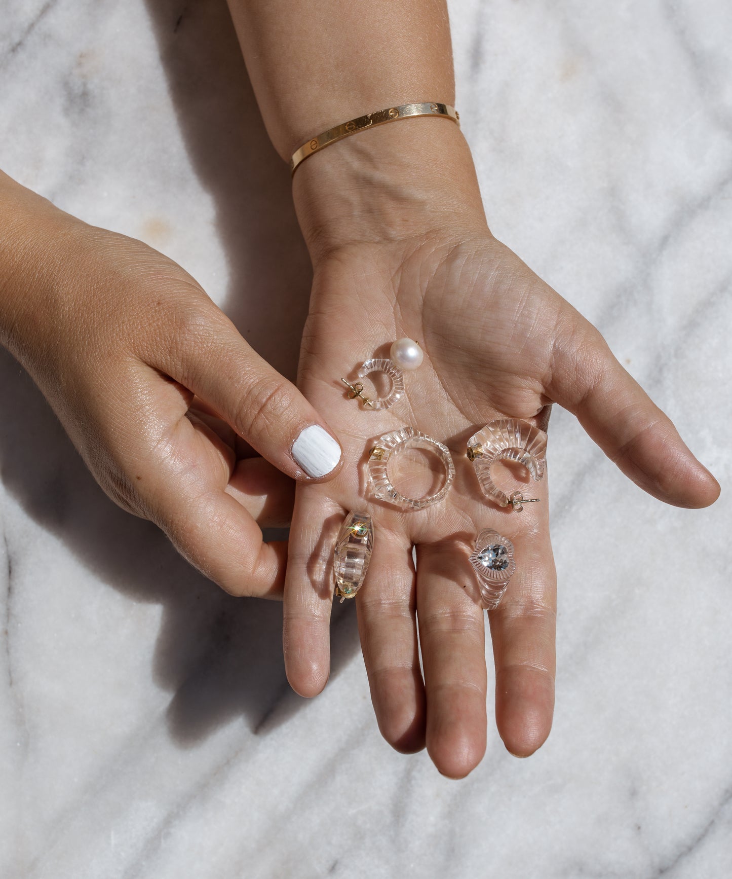 A hand elegantly displays several transparent and gold hoop earrings, shimmering like a WALD Berlin Be My Lover Transparent Ring, on a sleek marble surface.