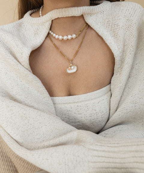 A person wears a cream sweater and top, accessorized with two necklaces: one delicate pearl strand and the WALD Berlin "Drop It Like It’s Hot Shell Necklace," featuring an exquisite gold-plated brass pendant.