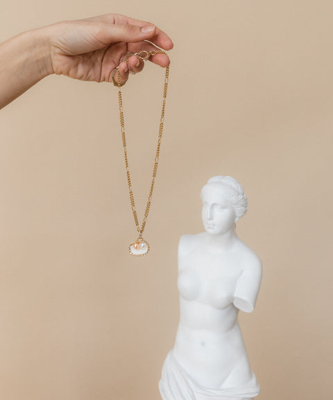 A hand displays the WALD Berlin "Drop It Like It's Hot" Shell Necklace in front of a small white bust sculpture of a woman, highlighting exquisite jewelry craftsmanship.
