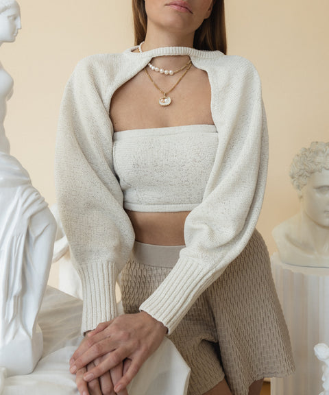 A person in a white knit shrug and tube top, adorned with WALD Berlin's Drop It Like It’s Hot Shell Necklace, stands near sculptures.