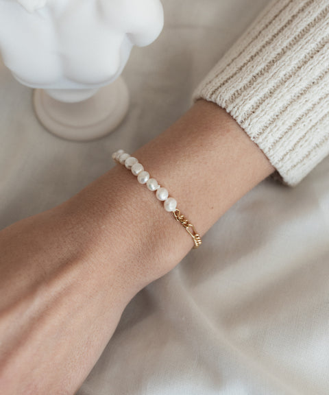 A person wearing the WALD Berlin Pas De Deux bracelet/anklet alongside a gold chain bracelet, with part of a cream-colored long sleeve visible.