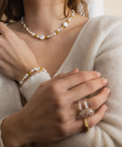 A woman elegantly displays her matching necklace, bracelet, and rings adorned with sweetwater pearls. She wears a light-colored sweater while showcasing the WALD Berlin Smilie Dude Pearl Bracelet with Extension, her hands gently crossed in front to complement the stunning jewelry.
