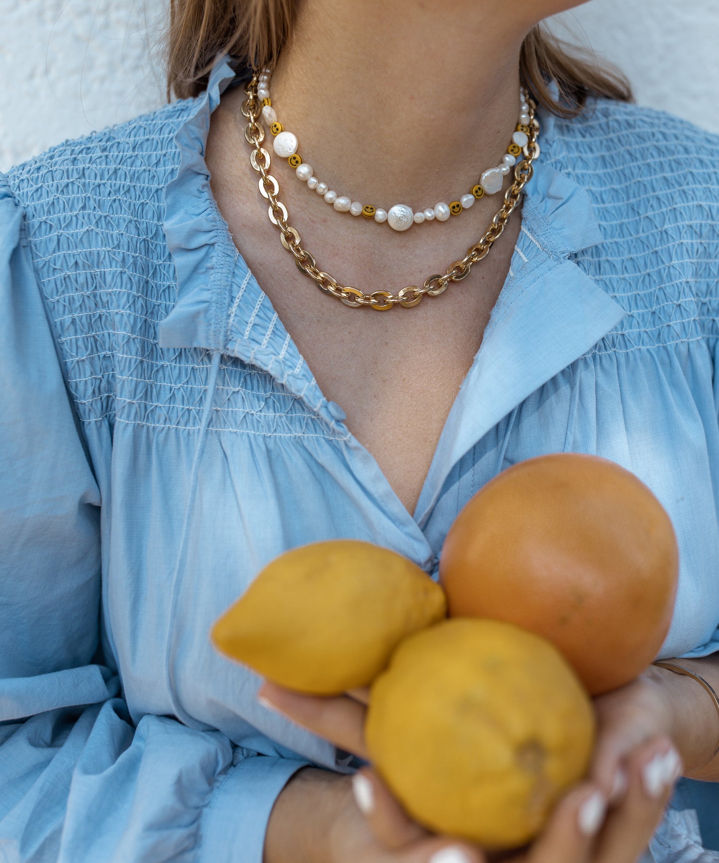 A person wears a blue blouse and is holding oranges and lemons while adorned with WALD Berlin's Smilie Dude Pearl Necklace featuring layered gold chains and smiley beads.