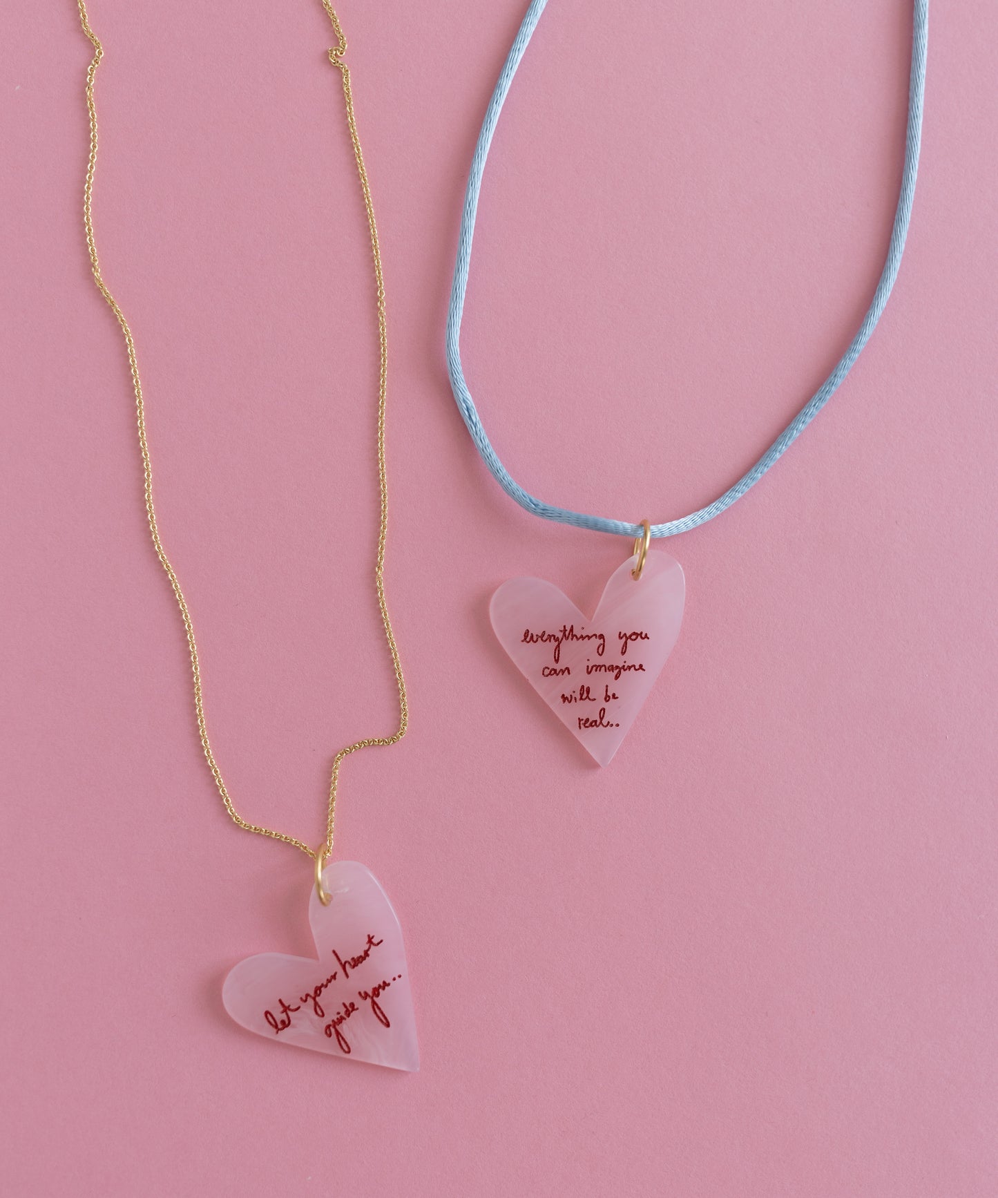 The "Let's be Happy Gold Necklace" by WALD Berlin features two heart-shaped pendants with inspirational text on an 18k gold-plated chain and blue cord, all against a pink background, inspiring positivity with every glance.