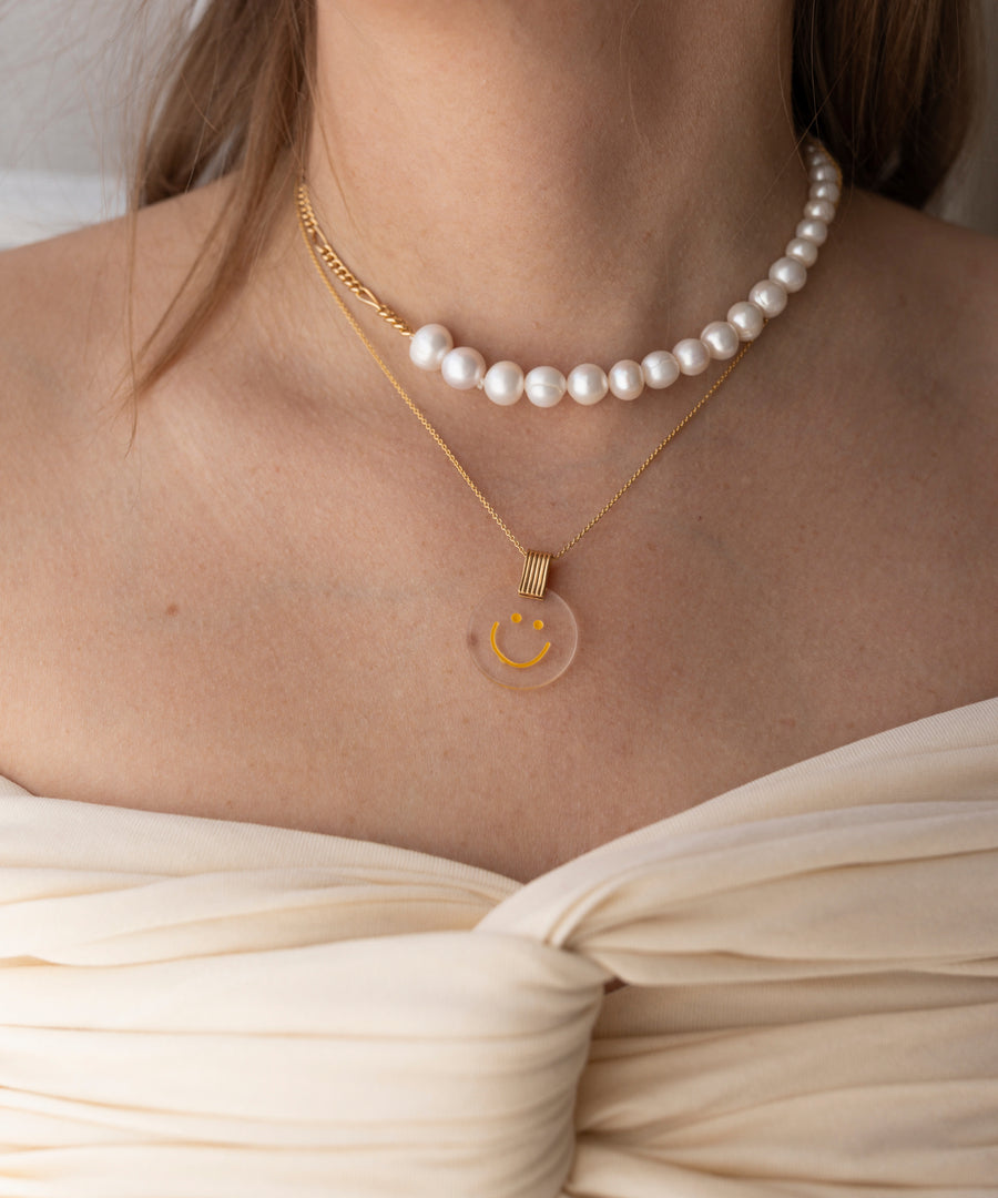 A woman wearing a white off-shoulder top, a pearl necklace, and the WALD Berlin Smilie Dude Medaillon Gold Necklace featuring a round yellow smiley face pendant, made in Germany.