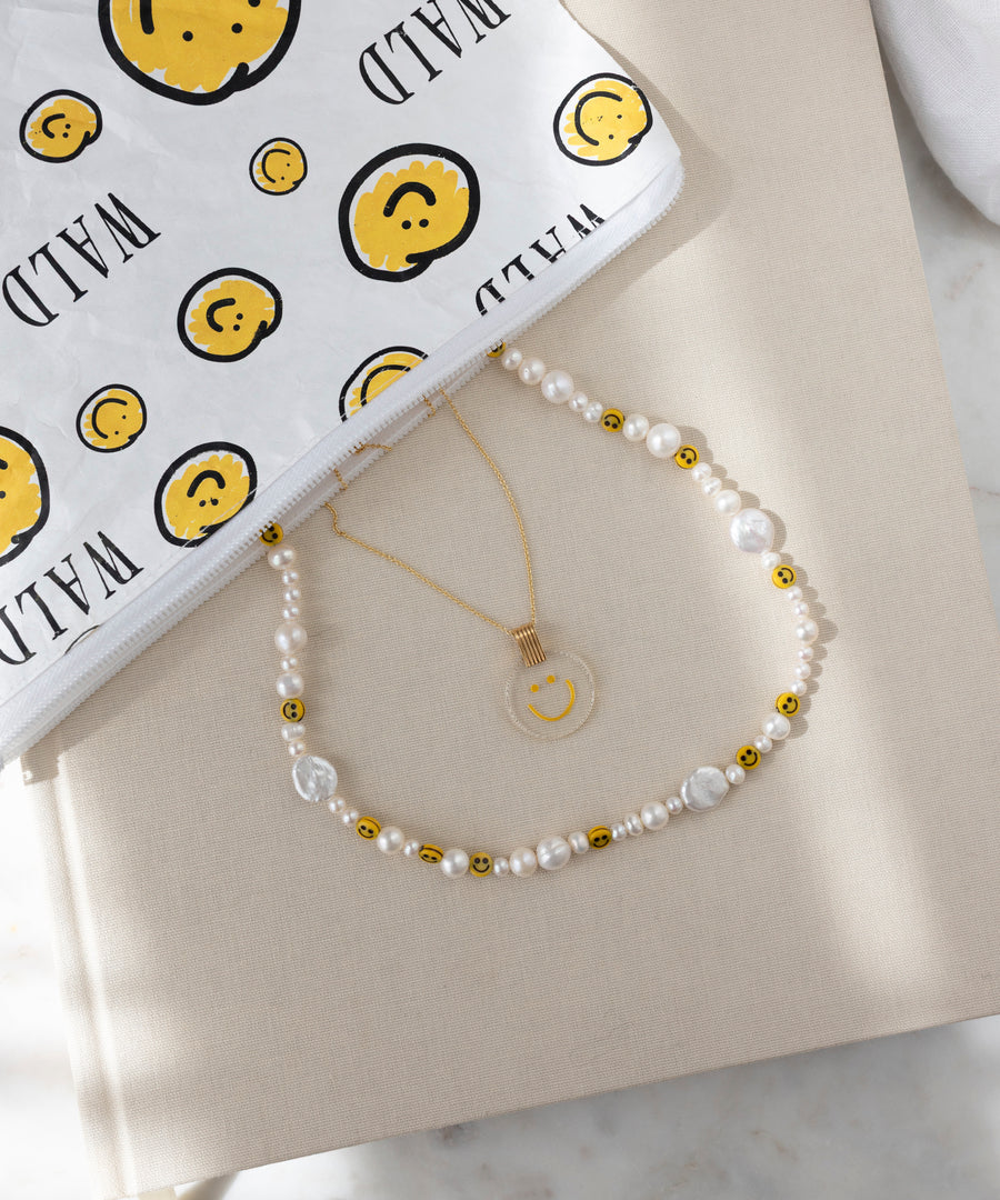 Die WALD Berlin Smilie Dude Perlenkette, handgefertigt aus Süßwasserperlen, Goldperlen und Smiley-Gesichtern, wird neben einer goldenen Anhängerkette und einem weißen Beutel mit gelben Smileys und dem Text "WILD" präsentiert.