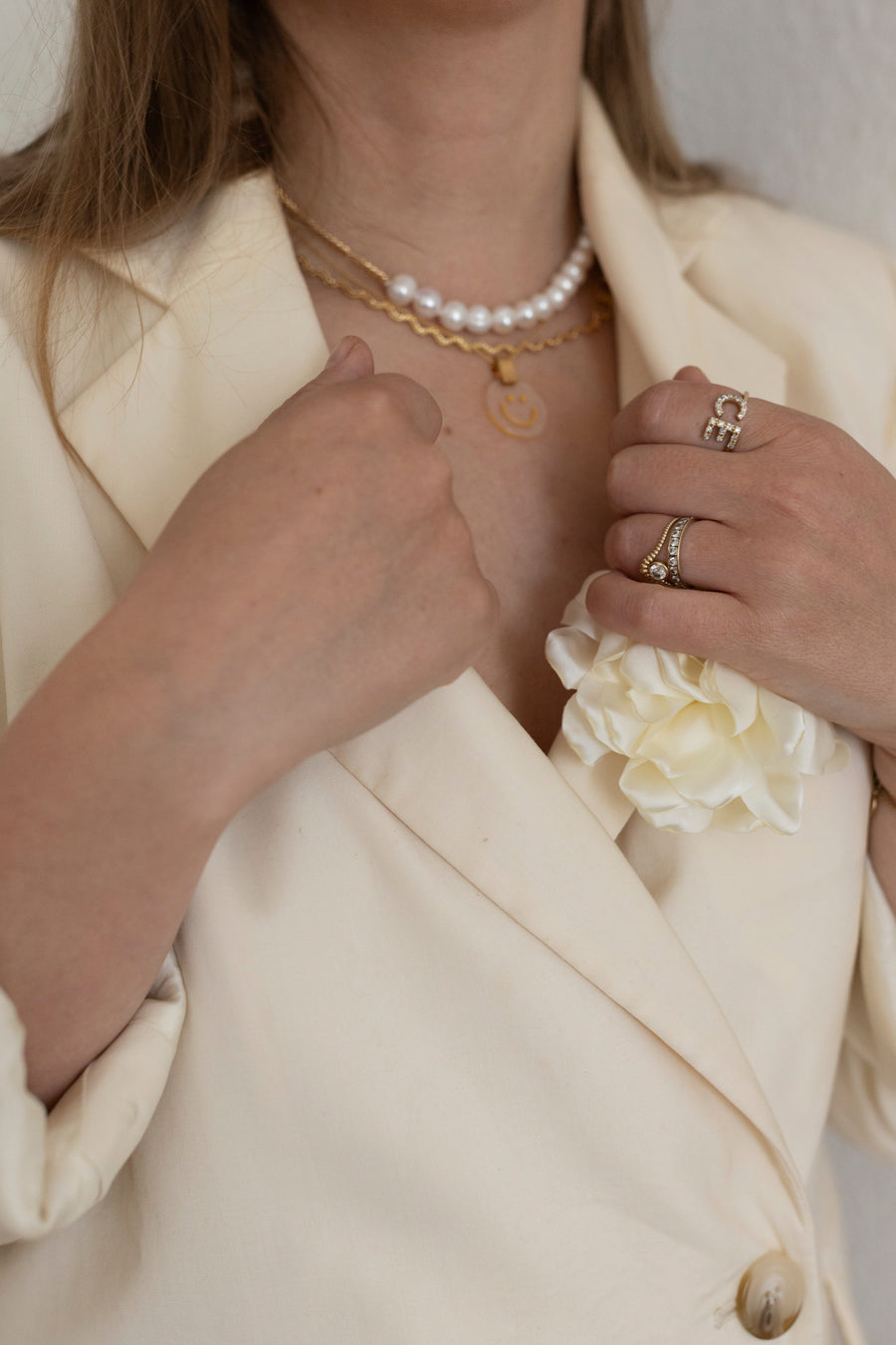 Eine Person trägt einen cremefarbenen Blazer mit einer weißen Reversblume, eine Perlenkette, eine WALD Berlin Irina Halskette Gold, Ringe an der linken Hand und ein Messingarmband.