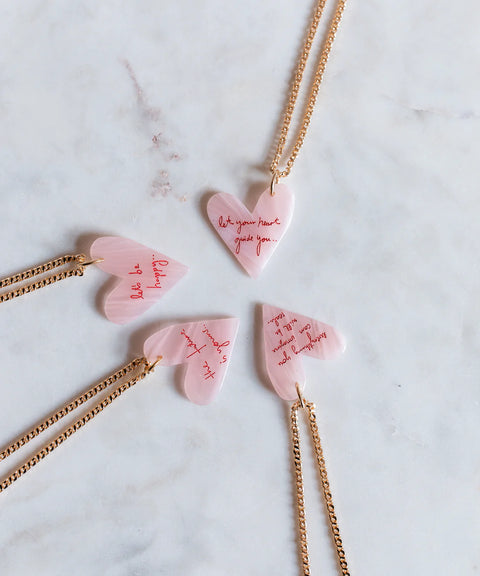 Vier rosafarbene herzförmige WALD Berlin "Let Your Heart Guide You"-Anhänger mit vergoldeten Ketten sind kreisförmig auf Marmor angeordnet und jeweils mit roter Kursivschrift graviert - eine ideale Halskette mit Herz oder ein persönliches Geschenk.