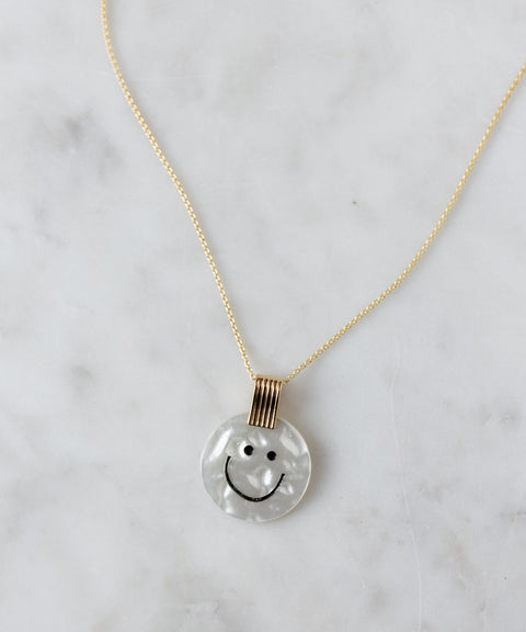 The WALD Berlin Smilie Dude Medaillon Pearl Necklace features an 18k gold-plated chain and a round white medallion with a black smiley face, perfect for adding playful elegance to any outfit.
