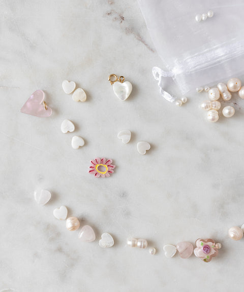 Assorted heart-shaped beads, a pink flower bead, and unique pearls from the Sweet Water Pearls Kit by WALD World are arranged on a marble surface. A sheer white drawstring pouch and more pearls from the DIY collection can be seen in the background.
