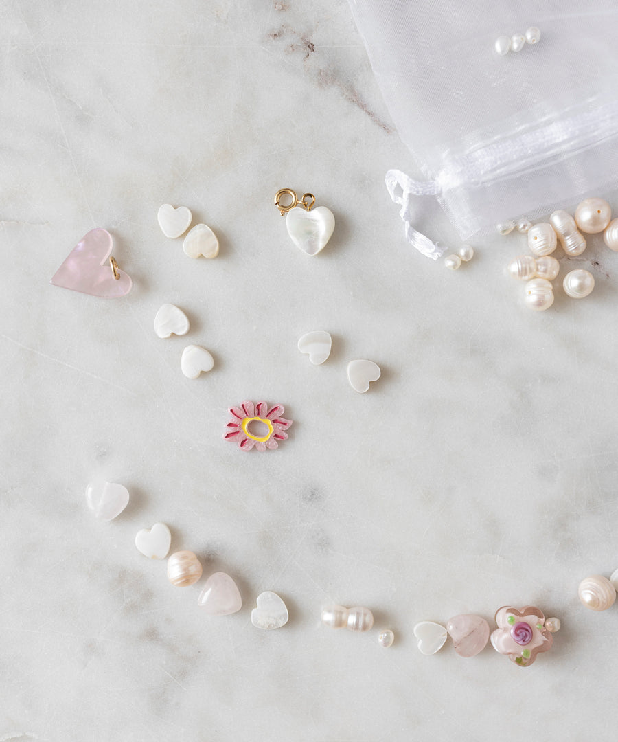 Assorted heart-shaped beads, a pink flower bead, and unique pearls from the Sweet Water Pearls Kit by WALD World are arranged on a marble surface. A sheer white drawstring pouch and more pearls from the DIY collection can be seen in the background.