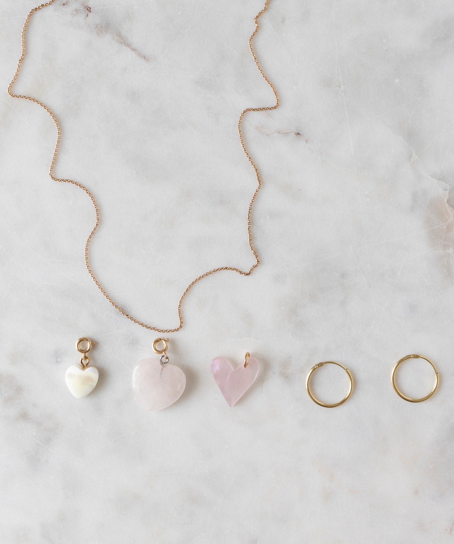 A Rose Quartz Heart Charm gold-plated brass necklace by WALD World, featuring three light rose, heart-shaped pendants, along with a pair of gold hoop earrings, are displayed on a marble surface.