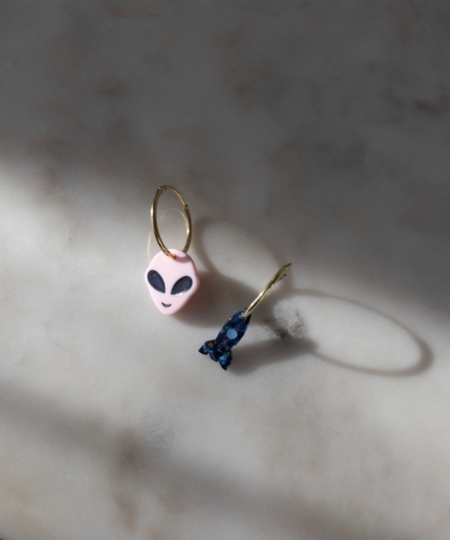 The SAMPLE SALE From Another Mother Gold-Blue Hoops by WALD Berlin are beautifully displayed on a marble surface; one earring is adorned with a pink alien head charm, while the other features a small dark blue figure. These earrings are crafted with precision and style using high-quality bio polyester elements, providing an eco-friendly touch and made in Germany.