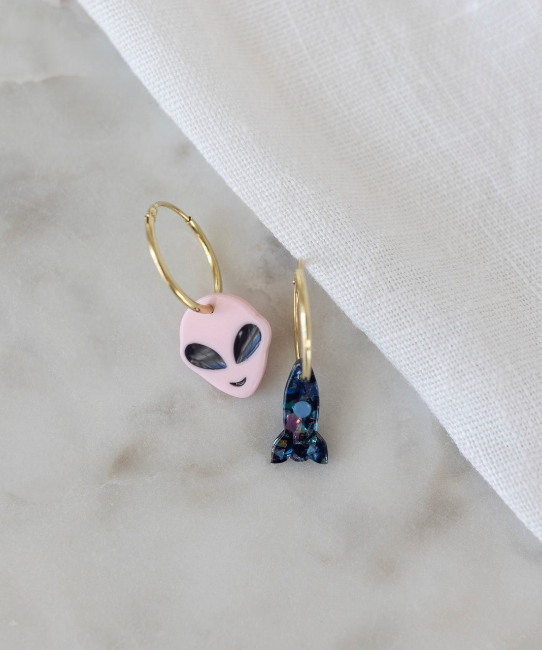 The SAMPLE SALE From Another Mother Gold-Blue Hoops from WALD Berlin feature a gold hoop earring set with pendants, one showcasing a pink alien head and the other adorned with a blue gemstone-encrusted rocket. Meticulously crafted in Germany, these earrings rest elegantly on a white marble surface partially draped with a white cloth, highlighting their exceptional craftsmanship.