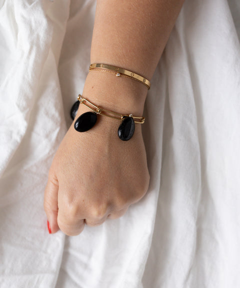 A hand with a gold bangle and the WALD Berlin Collection Noir Ashley Drop Gold Bracelet featuring three black stone charms rests on white fabric.