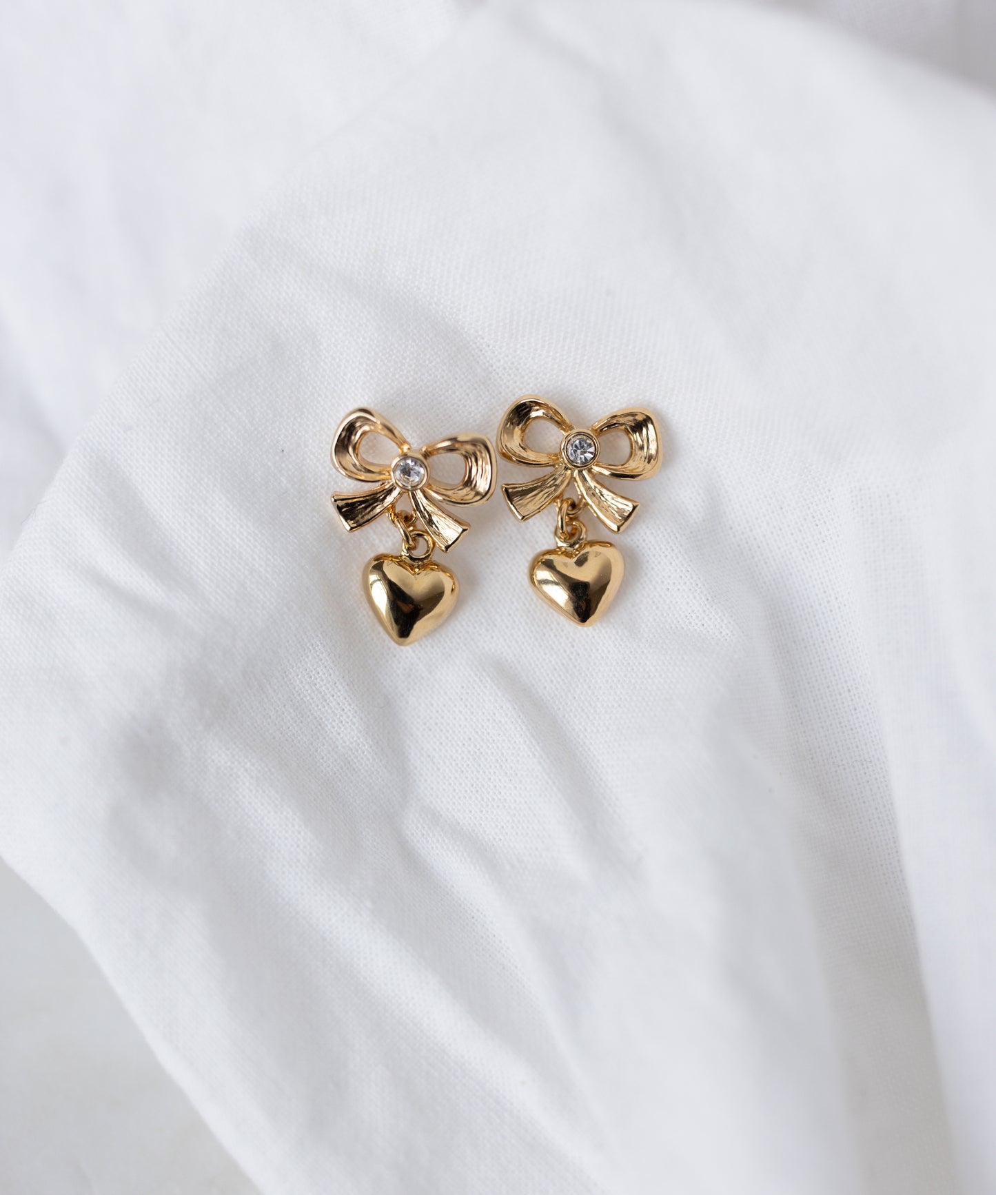 WALD Berlin's Valentines Day special bow heart earrings—gold-plated brass with bow designs and dangling heart charms—are shown on a white fabric background. Perfect as heart earrings for Valentine's Day.
