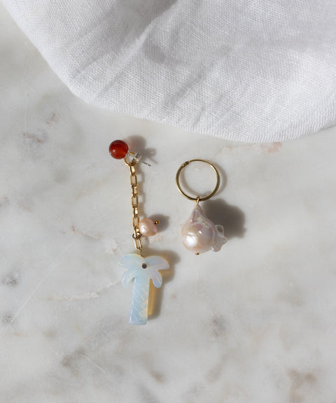 The Mexicana Mariachi Earrings Pearl by WALD Berlin are mismatched gold earrings with pearls, red bead, chain, and palm tree charm, displayed on marble. Handmade in Germany. A piece of white fabric is visible in the corner.
