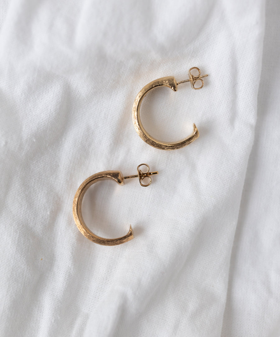 The SAMPLE SALE Pop Classy Hoop Gold earrings by WALD Berlin, featuring 18k gold-plated textured surfaces, are displayed on a white fabric background.