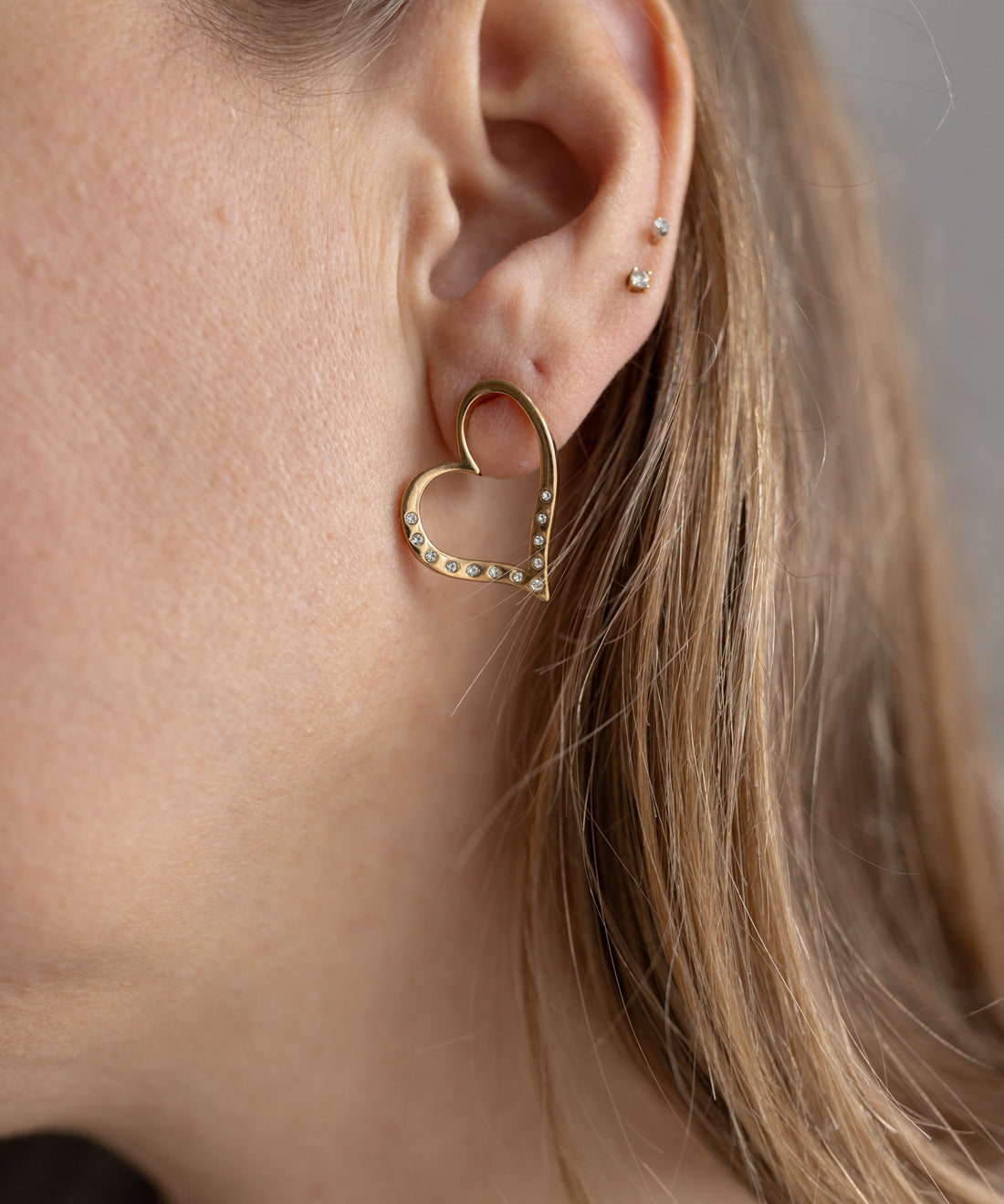 Close-up of a person's ear wearing WALD Berlin's SAMPLE SALE Valentines Day special sketch heart earrings, goldplated and gemstone-studded; three small studs are also visible.