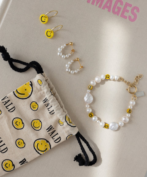 A fabric pouch adorned with smiley faces holds WALD Berlin’s Smilie Yellow Earrings, pearl earrings, a pearl bracelet, and smiley face stud earrings on a beige surface with partially visible pink text. The collection features elegant 18k gold-plated hoops for a luxurious accent.