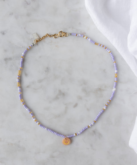 The Pop Smilie Mini necklace by WALD Berlin, made with light purple and 18k gold plated beads and a small round Smilie Medaillon pendant, is shown on marble beside a white cloth.