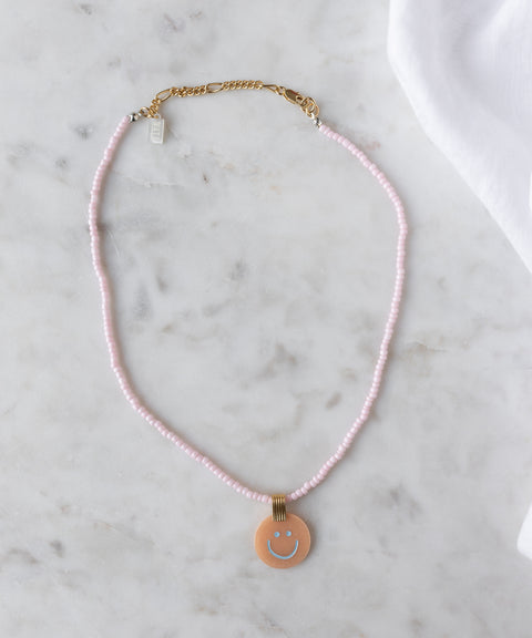 The Pop Smilie Medallion Peach by WALD Berlin is a pink beaded necklace with a gold clasp and round pendant featuring a blue smiley face—ideal for adding playful charm to classic styles or your summer look.