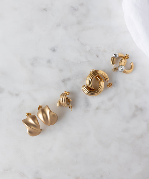 Four pairs of 18k gold plated earrings, including hoops and abstract shapes, from WALD Berlin's SAMPLE SALE Matt Gold Creoles collection are arranged on white marble—exclusive picks from our sample sale.