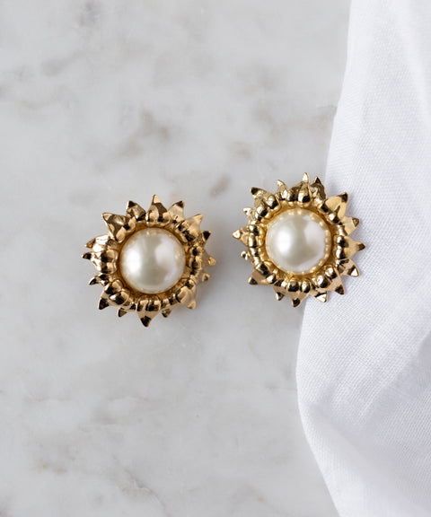 The SAMPLE SALE Pop Soleil Earrings by WALD Berlin feature gold plating, pearl studs, and a textured spiked border, shown on white marble beside soft fabric—a striking statement jewelry piece.