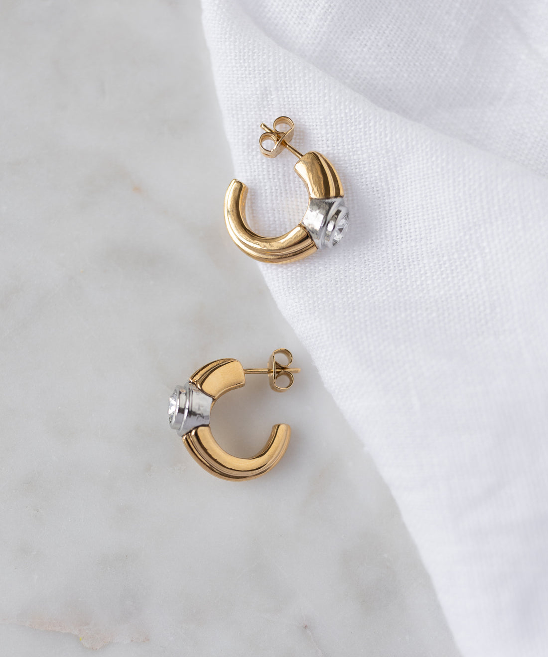 The SAMPLE SALE Pop Bi Color Crystal Earrings by WALD Berlin, with gold and silver tones, are displayed on a white marble surface partially covered by a white cloth.