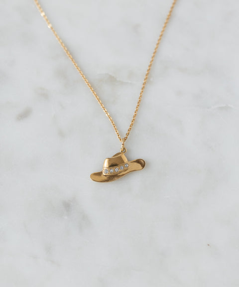 The SAMPLE SALE Pop Cowgirl Necklace by WALD Berlin is a gold necklace from Germany with a 55cm chain, featuring a cowboy hat-shaped pendant with clear gemstones, shown on white marble.