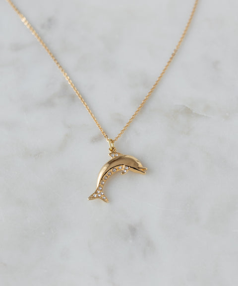 The WALD Berlin Pop Dolphin Necklace, a goldplated brass piece with a dolphin-shaped pendant embellished with sparkling Swarovski stones, is elegantly displayed on a light marble surface.