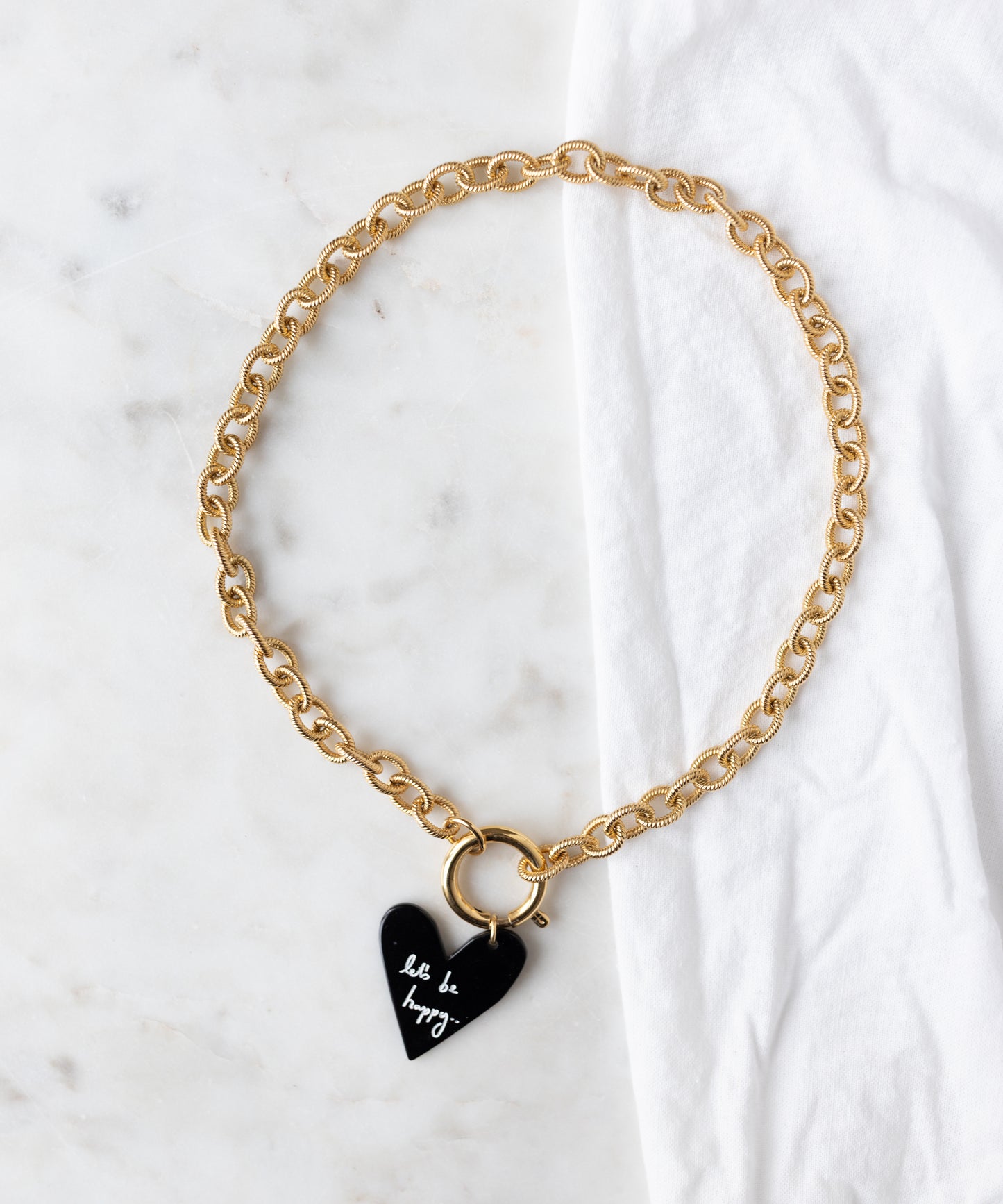 The WALD Berlin Collection Noir Let’s Be Happy Gold Necklace features a black heart-shaped pendant engraved with "let it be, happy," and 18k gold plated details, shown on a white marble surface beside a white cloth.