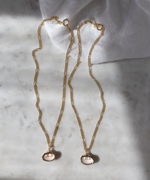 The Drop It Like It’s Hot Shell Necklace set by WALD Berlin features two handmade gold-plated chains with oval pearl pendants, elegantly displayed on marble near a white cloth, radiating timeless charm.